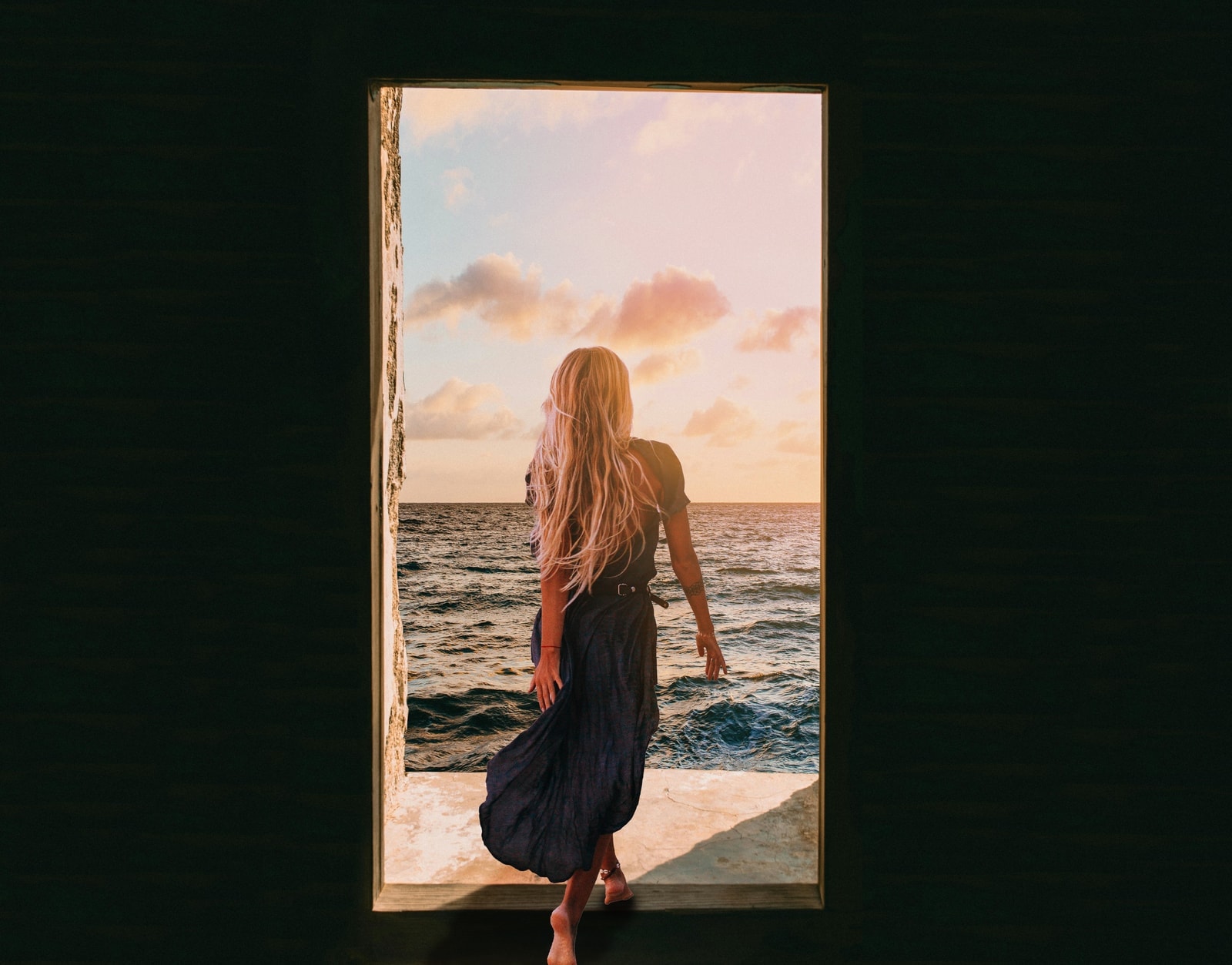 Photo manipulation composite — a woman in a doorway looking out over the ocean at sunset.